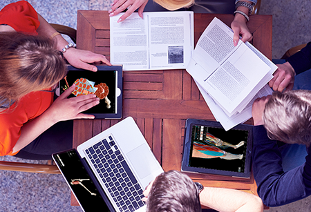 4 estudiantes estudiando alrededor de una mesa con libros de texto abiertos, 2 tabletas y un ordenador portátil, todos mostrando la anatomía en 3D de Visible Body en las pantallas